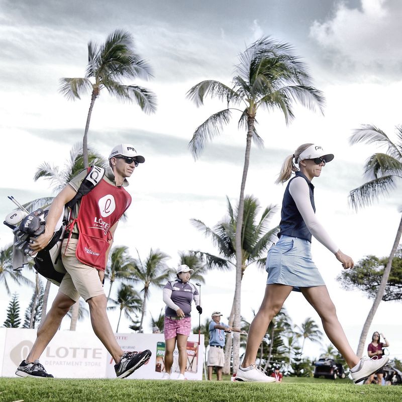 Daniel Taylor carries Pernilla’s golf bag as they walk down a fairway lined with palm trees